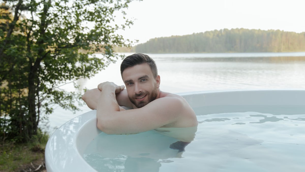 Shirtless man enjoying relaxation in hot tub by scenic lake, smiling and embracing leisure outdoors.
